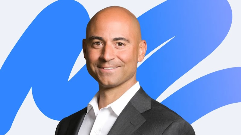 Damon Berger, SVP of marketing shared services, Gap Inc., wearing a crisp white shirt and black suit jacket, smiles warmly, against a white background with a large blue squiggle shape