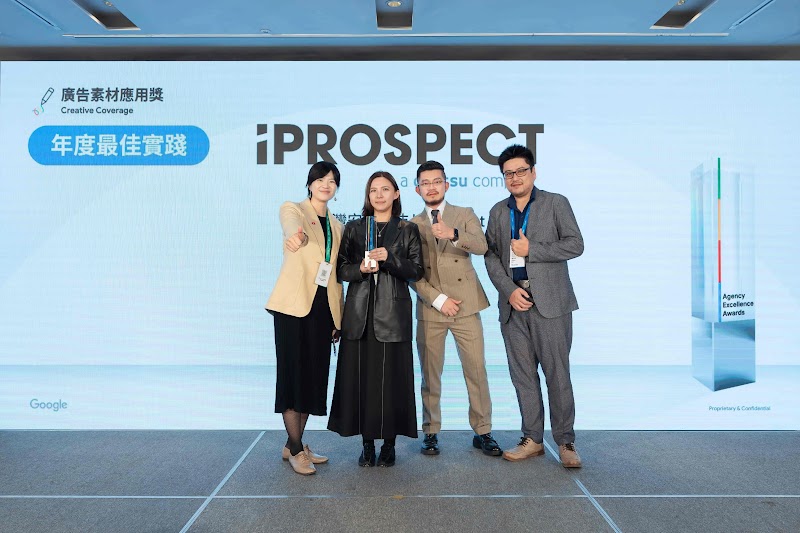 iProspect團隊四位成員於Google頒獎典禮舞台上，背景展示「iProspect」標誌及「廣告素材：年度最佳實踐」藍色獎徽。左起第一位女性著淡黃色西裝外套、黑色褲裝；第二位女性著黑色長版外套、黑色褲裝；第三位男性著米色西裝；第四位男性著灰色西裝。所有成員皆配戴Google Agency Excellence Award證件牌，呈現專業祝賀姿態。此為iProspect展示YouTube全漏斗策略與AI驅動的廣告素材創意成果。