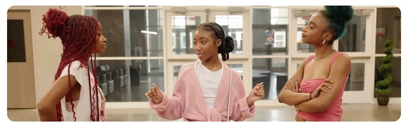 Three Black teen girls talk while standing in a wide hall with interior windows behind them. Their clothing is trendy and youthful, in pastel pink, peach, and white.
