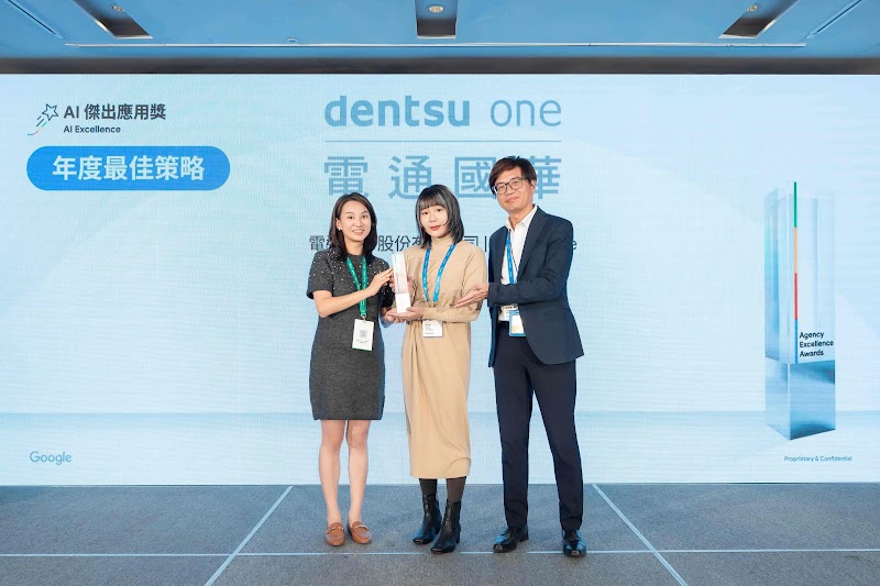 電通國華團隊三位成員於Google頒獎典禮舞台上慶祝。左側女性著深灰色短袖洋裝、棕色皮鞋，中央女性著淡黃色長版外套、黑色短靴，右側男性著深藍色西裝。三人皆配戴藍色證件帶。背景展示「dentsu one」標誌與「AI傑出應用獎：年度最佳策略」藍色獎徽。團隊以Google AI驅動的全漏斗行銷策略，在兩月內達成122倍新客名單成長，贏得2025 Google Award肯定。
