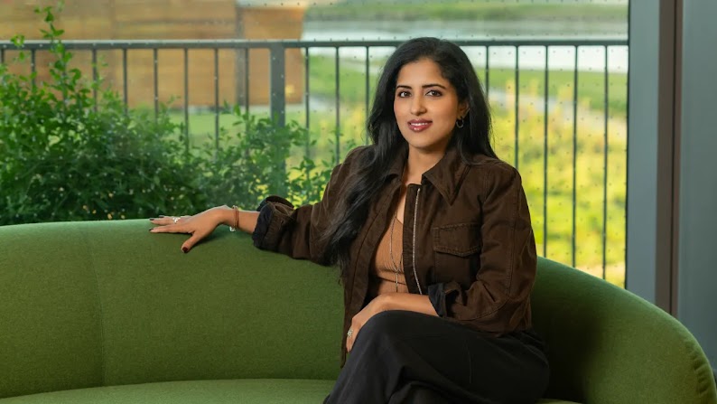 Vidhya Srinivasan, VP und General Manager of Ads and Commerce bei Google, sitzt auf einer grünen Couch vor einem Fenster mit Blick auf einen Strauch und ein Terrassengeländer. Srinivasan hat langes dunkles Haar und trägt eine braune Jacke und eine schwarze Hose.