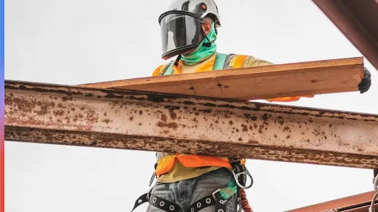 An electrician in a helmet and face shield rests a wooden beam on top of a metal beam