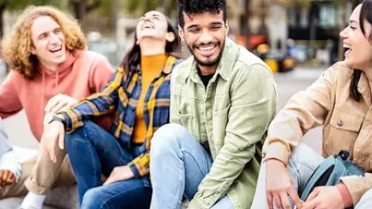 A group of four diverse young adults sitting outdoors on stone steps, laughing and talking together in a bright, urban setting. They are dressed in casual autumnal clothing, symbolizing the vibrant and connected younger generation in Europe.