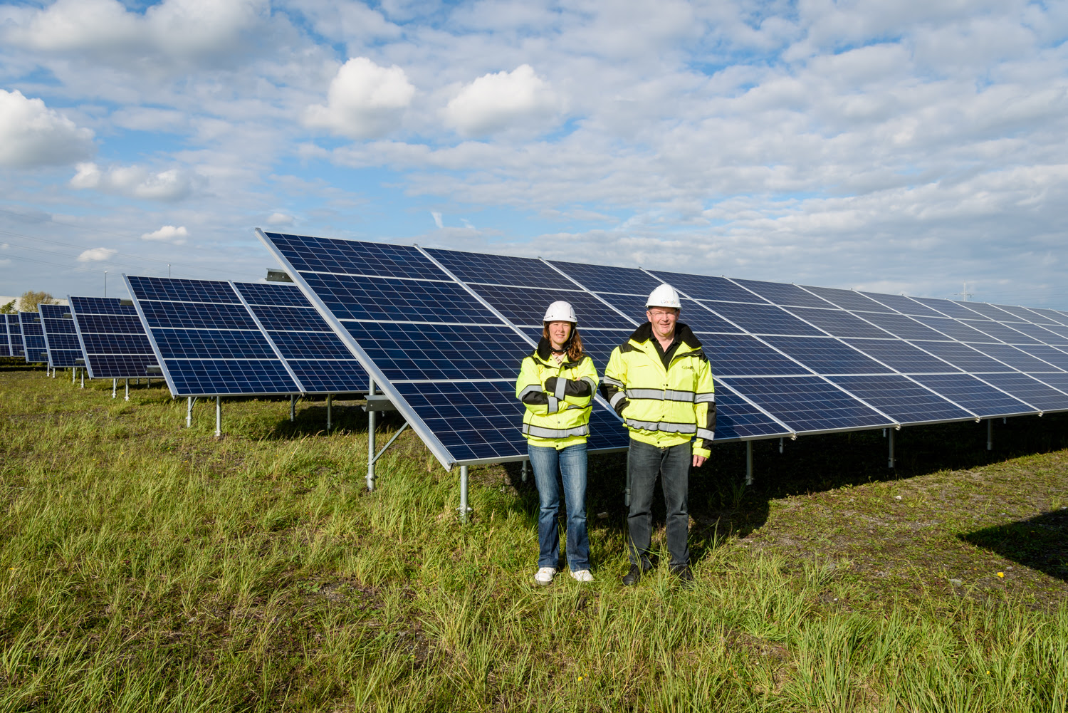 Two people stand in front of large solar panels in a field.