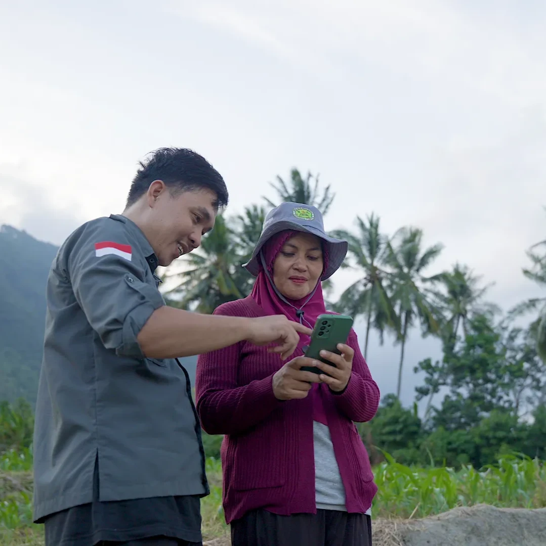 A man and a woman wearing a hat look down at a phone, smiling. They are in a rural area, with palm trees in the background.