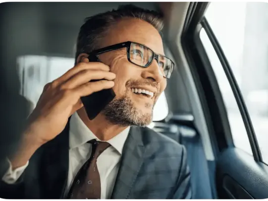 A white man wearing a gray checkered suit and burgundy tie talking on his mobile phone while looking out the window in the backseat of a car.