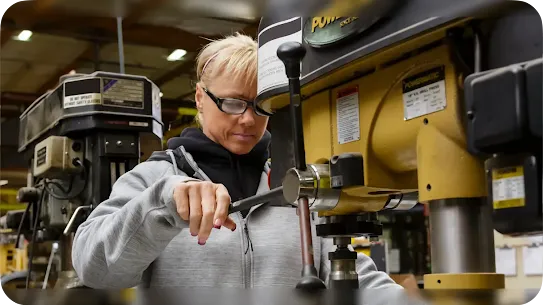 A woman wearing safety glasses and a grey hoodie operates a heavy-duty yellow Powermatic drill press in a workshop. She is focused, with her hand on the machine's lever.