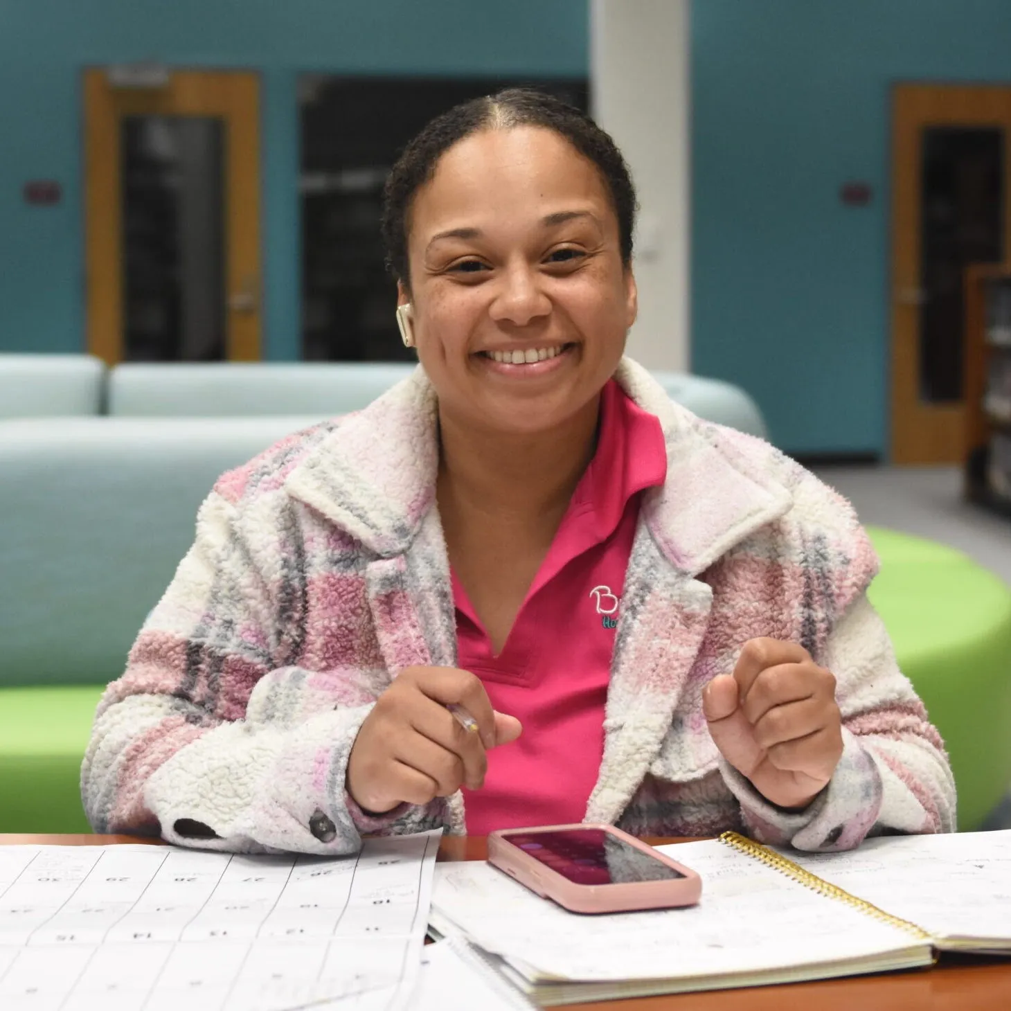a woman sits at a desk with papers at the library. She smiles at the camera.