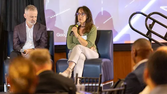 A panel discussion taking place on a stage at an indoor event. A woman with long dark hair, wearing glasses, a green long-sleeve top, and light-colored pants, sits with her legs crossed on a dark blue leather armchair while speaking into a microphone. Seated to her left on an identical chair is a man with short grey hair and a beard, wearing a dark plaid blazer and a white button-down shirt, looking downward. An audience is visible in the blurred foreground, and a large projection screen displays a bright abstract graphic behind the speakers.