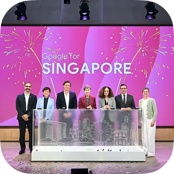 A diverse group of seven professionals standing behind a glass display featuring the Singapore skyline outline. The background is a large pink screen with the text "Google for SINGAPORE" and firework graphics.