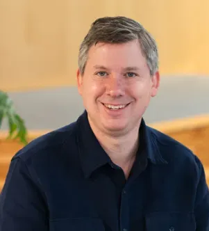 A professional portrait of a smiling man with short salt-and-pepper hair wearing a navy blue button-down shirt, featured in a case study for Julius Education in Massachusetts.