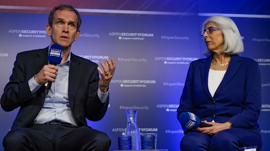 A man and a woman speaking on a panel at the Aspen Security Forum. The man on the left, wearing a dark suit and light blue shirt, is speaking into a microphone and gesturing with his hand. The woman on the right, in a blue blazer and glasses, listens attentively while holding a microphone. The background features a blue wall with the "Aspen Security Forum" and "Aspen Institute" logos repeated.