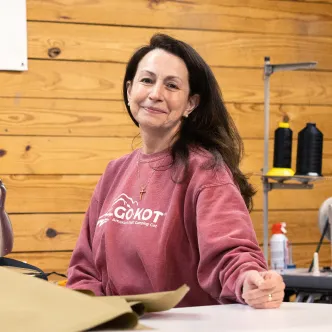 A woman wearing a pink Go-Kot sweatshirt smiles in a workshop setting with a wooden wall and spools of black thread in the background.