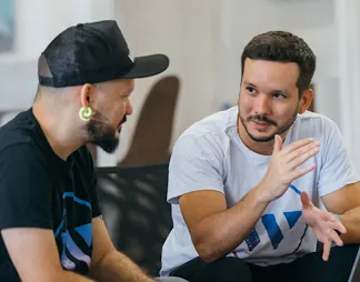 A man in focus is discussing with other man wearing black cap while sitting in front of laptop