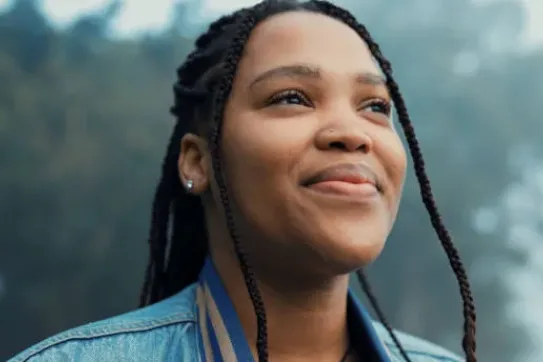 A smiling young woman with braided hair wearing a denim jacket, looking upward in an outdoor setting.