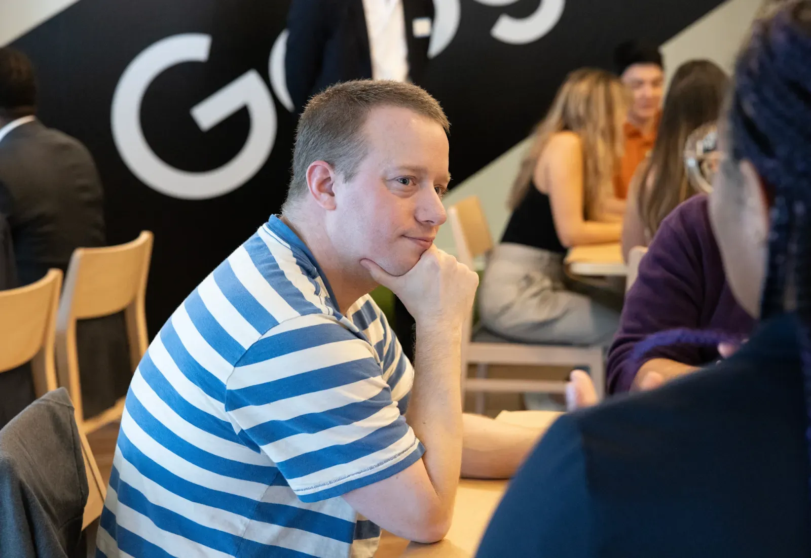 Nathan Havens, a man in a blue and white striped t-shirt, sits at a wooden table with his hand on his chin, listening intently during a discussion. In the background, a large "Google" logo is partially visible on a black wall, along with other participants in a bright office setting.