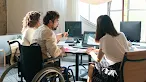 Three colleagues, including a man seated in a wheelchair who is pointing at a monitor, collaborate around a desk in a modern office.