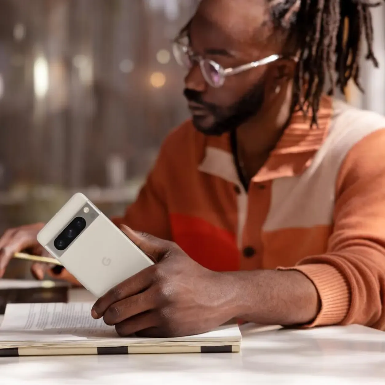 A man with long dark dreadlocks, a beard, and glasses is seated at a table, looking down at an open book. He is holding a white smartphone in his right hand, showcasing its camera bar with three lenses. He wears an orange and cream color-blocked knit cardigan. The background is softly blurred with warm, ambient lighting.