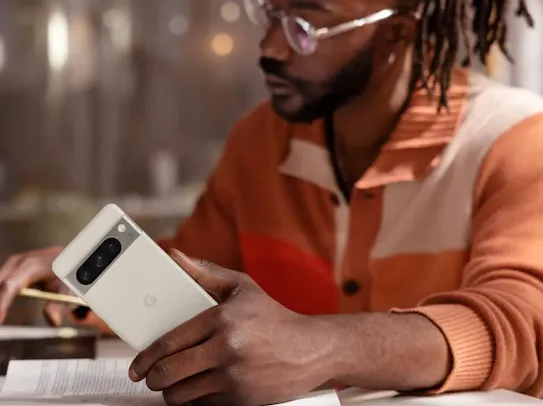 A man with long dark dreadlocks, a beard, and glasses is seated at a table, looking down at an open book. He is holding a white smartphone in his right hand, showcasing its camera bar with three lenses. He wears an orange and cream color-blocked knit cardigan. The background is softly blurred with warm, ambient lighting.