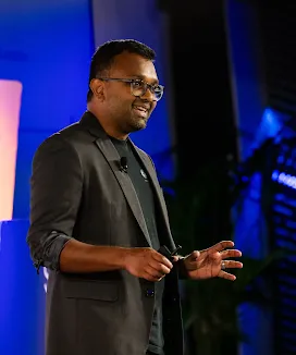 A man on the event stage is addressing the audience while holding microphones.