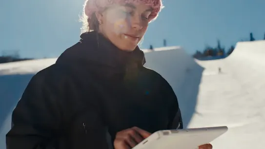 A Team USA winter athlete in a black jacket and pink beanie stands on a snowy halfpipe under a bright sun. She is looking down and interacting with a white tablet, using Google Cloud AI tools to analyze performance data and elevate her tricks during training.