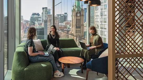 Three people sit in a sunlit office lounge, talking on a green sofa by a large window overlooking a cityscape. Two are using laptops.
