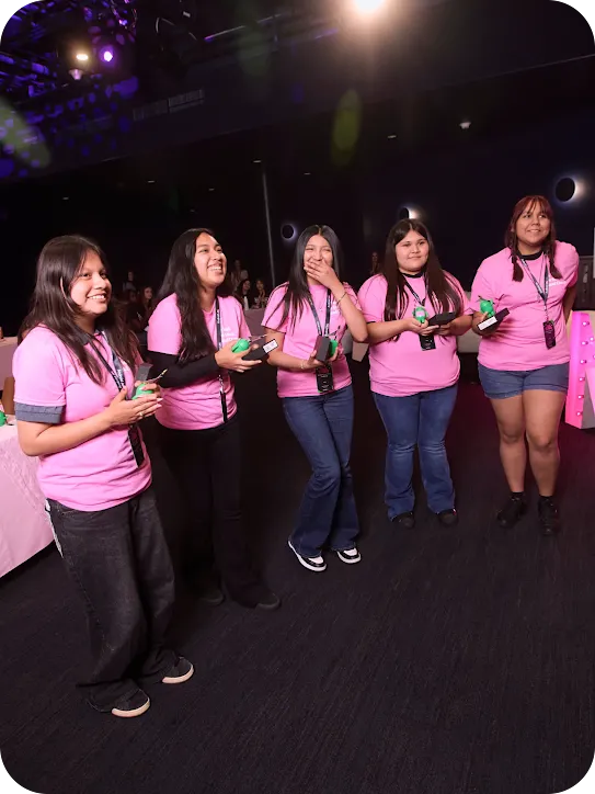 Five girls in pink shirts standing together on a stage laughing; some are holding smartphones and small Android mascots.
