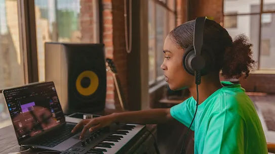 Young Black woman wearing over-ear headphones producing music at a desk with a laptop, MIDI keyboard, and studio monitor in a loft-style room.