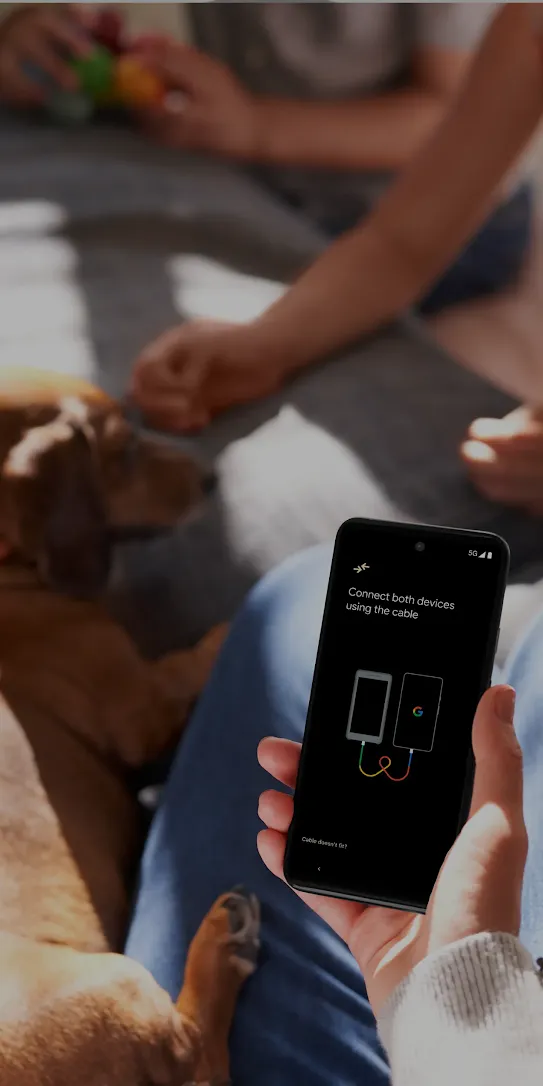A person is holding an Android phone while petting a brown dog. The phone screen has a picture of two devices connected by a cord. Another person’s hands close to the dog’s face is in the background.