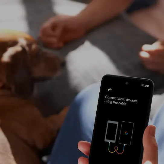 A person is holding an Android phone while petting a brown dog. The phone screen has a picture of two devices connected by a cord. Another person’s hands close to the dog’s face is in the background.