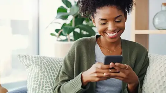 A smiling woman with curly hair sits comfortably on a sofa, holding and looking down at a smartphone in her hands.