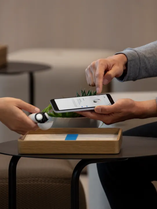 Eine Hand hält Pixel Buds in der Farbe Porcelain über einen braunen Tisch. Eine andere Hand zeigt auf ein Smartphone-Display.