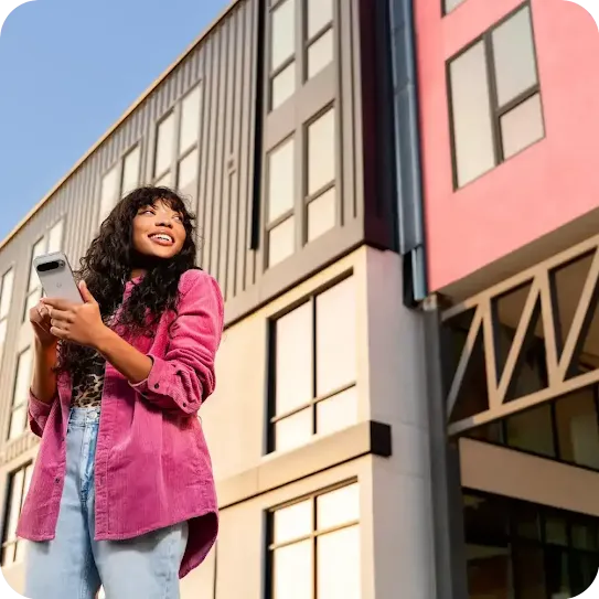 A low-angle view of a young woman holding her phone and smiling in front of a modern urban building.