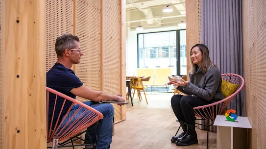 Two colleagues sit facing each other in pink cord chairs within a cozy, semi-enclosed meeting nook made of light-colored wood with perforated pegboard walls. On the right, a woman in a grey turtleneck and black boots gestures with her hands while speaking. Opposite her, a man in a navy t-shirt and jeans listens attentively with a closed laptop on his lap. A small white side table next to the woman holds a colorful "G" logo. In the blurred background, a bright, open-plan office space with yellow chairs and large windows is visible.
