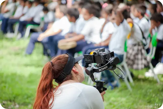 An over-the-shoulder view of a woman with auburn hair in a ponytail and a black visor, holding a professional camera on a DJI Ronin gimbal. She is filming a crowd of school children sitting in rows on green grass in the blurred background.