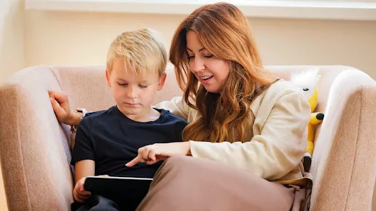 A woman and her son are sitting in an armchair looking at a tablet together while she points at the screen.