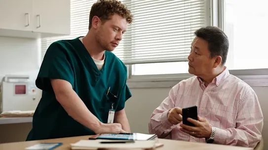 A male nurse in green scrubs leans over a table to consult with an older Asian man in a pink checkered shirt. The patient is holding a smartphone while the nurse points to a tablet on the desk in a bright medical office setting.