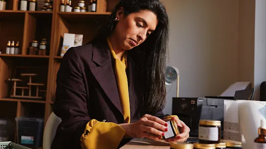 A dark-haired woman, wearing a jacket over a mustard blouse, inspects a small jar while sitting at a desk with more products.