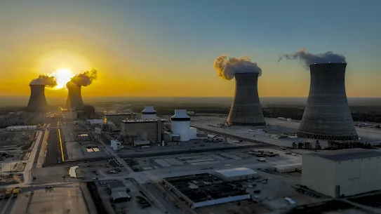 An aerial wide-angle shot of the Vogtle Electric Generating Plant in Georgia at sunset, featuring four massive cooling towers with steam rising against a golden horizon and the industrial complex in the foreground.