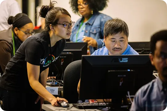 A female instructor named Erika wearing glasses leans over a desk to guide a man sitting in front of a computer monitor in a busy classroom setting.