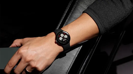 A wrist wearing a black Google Pixel Watch resting on a dark surface.
