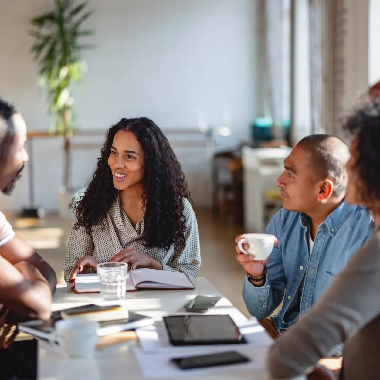 Four people are gathered around a table in a bright, modern office space, engaged in a conversation. A woman with long curly hair and a patterned shirt is smiling as she looks at a man across the table. Another man in a blue denim shirt holds a white mug and listens intently. The table is covered with notebooks, tablets, smartphones, and glasses of water, with soft sunlight casting shadows across the surface.