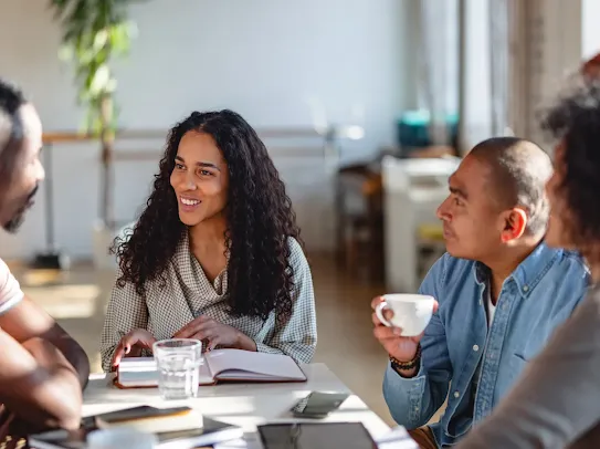 Four people are gathered around a table in a bright, modern office space, engaged in a conversation. A woman with long curly hair and a patterned shirt is smiling as she looks at a man across the table. Another man in a blue denim shirt holds a white mug and listens intently. The table is covered with notebooks, tablets, smartphones, and glasses of water, with soft sunlight casting shadows across the surface.