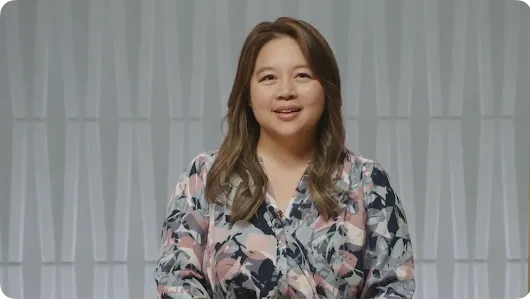 A medium close-up shot of a woman with long, wavy brown hair, smiling and looking directly into the camera. She is wearing a patterned pink, blue, and white floral-style blouse with a small lavalier microphone attached, standing in front of a modern, light-gray geometric textured wall.
