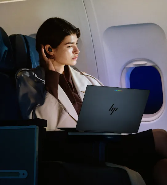 A person working on an HP laptop while sitting in a business class seat on an airplane at night. They are looking out the window, and the cabin is dimly lit.