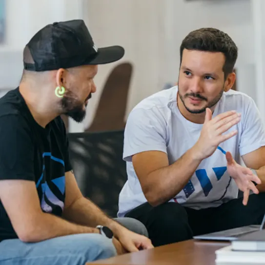 A man in focus is discussing with other man wearing black cap while sitting in front of laptop.
