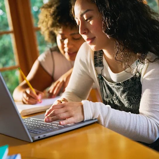 Two people at a desk, one looking at a laptop, the other writing in a notebook.