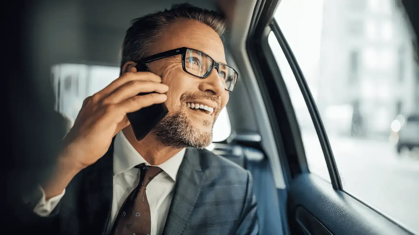 A white man wearing a gray checkered suit and burgundy tie talking on his mobile phone while looking out the window in the backseat of a car.