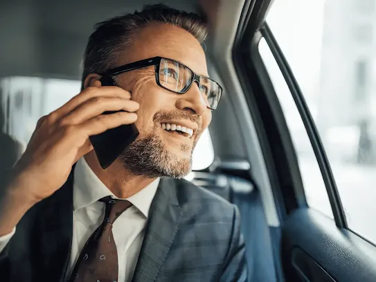 A white man wearing a gray checkered suit and burgundy tie talking on his mobile phone while looking out the window in the backseat of a car.