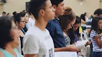 A crowd of people standing in a hall, focusing on a young woman in the foreground reading from a book.